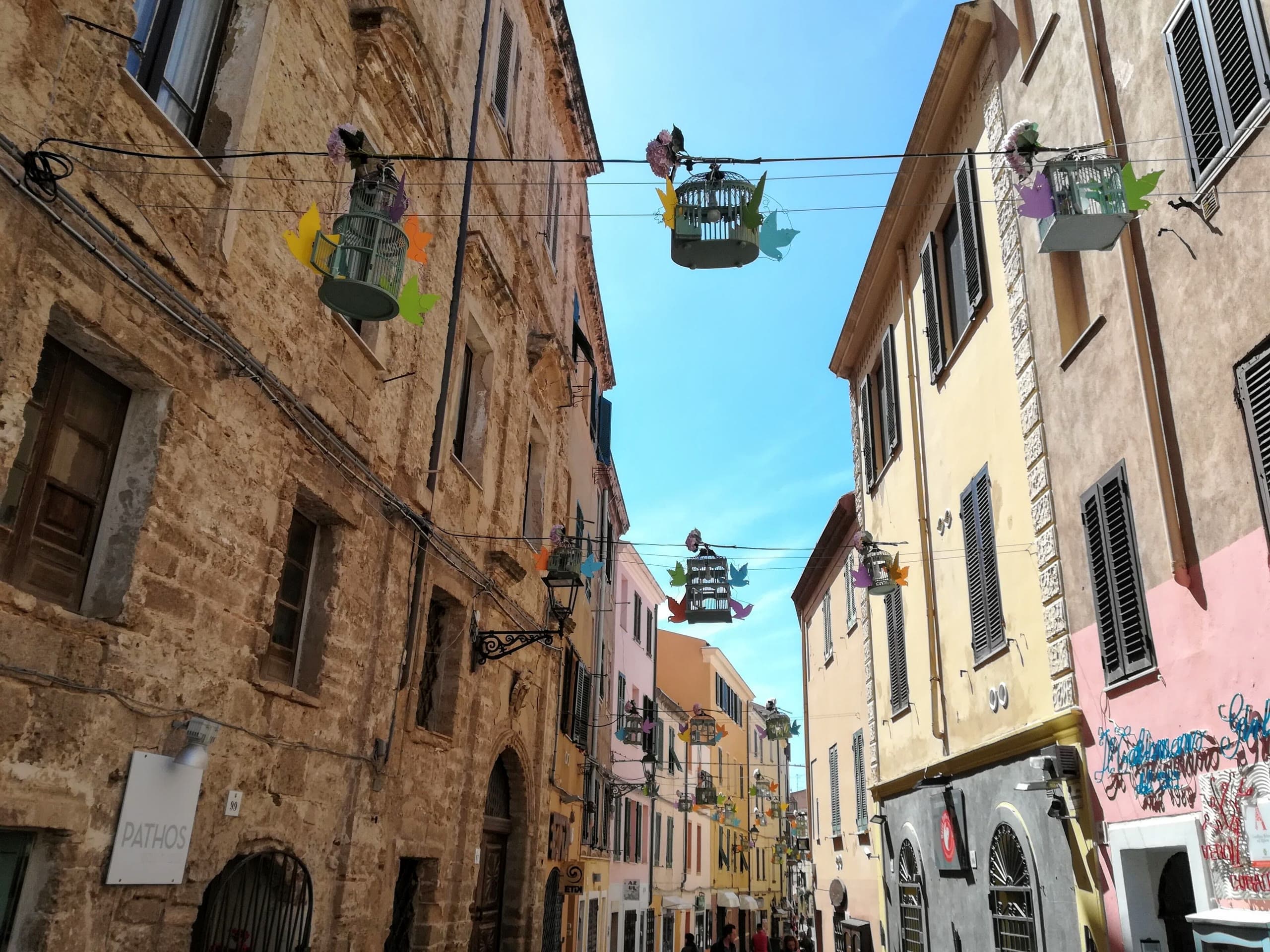 A narrow lane in Alghero's old town lined with sandstone and pastel facades, decorated birdcages hanging on wires overhead
