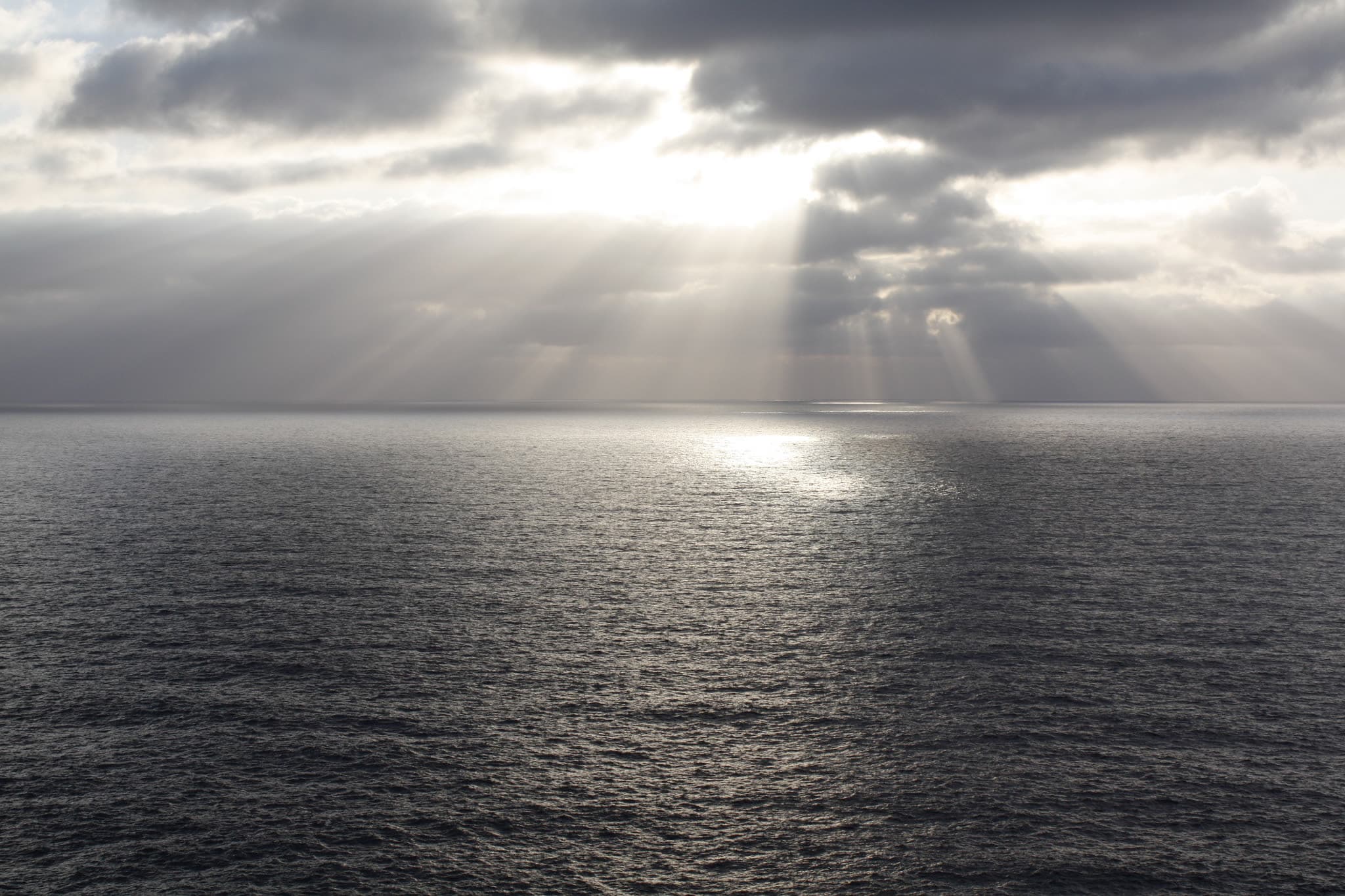 Sunbeams breaking through a deck of cloud over open sea off Alghero, the water dark and rippled below the horizon