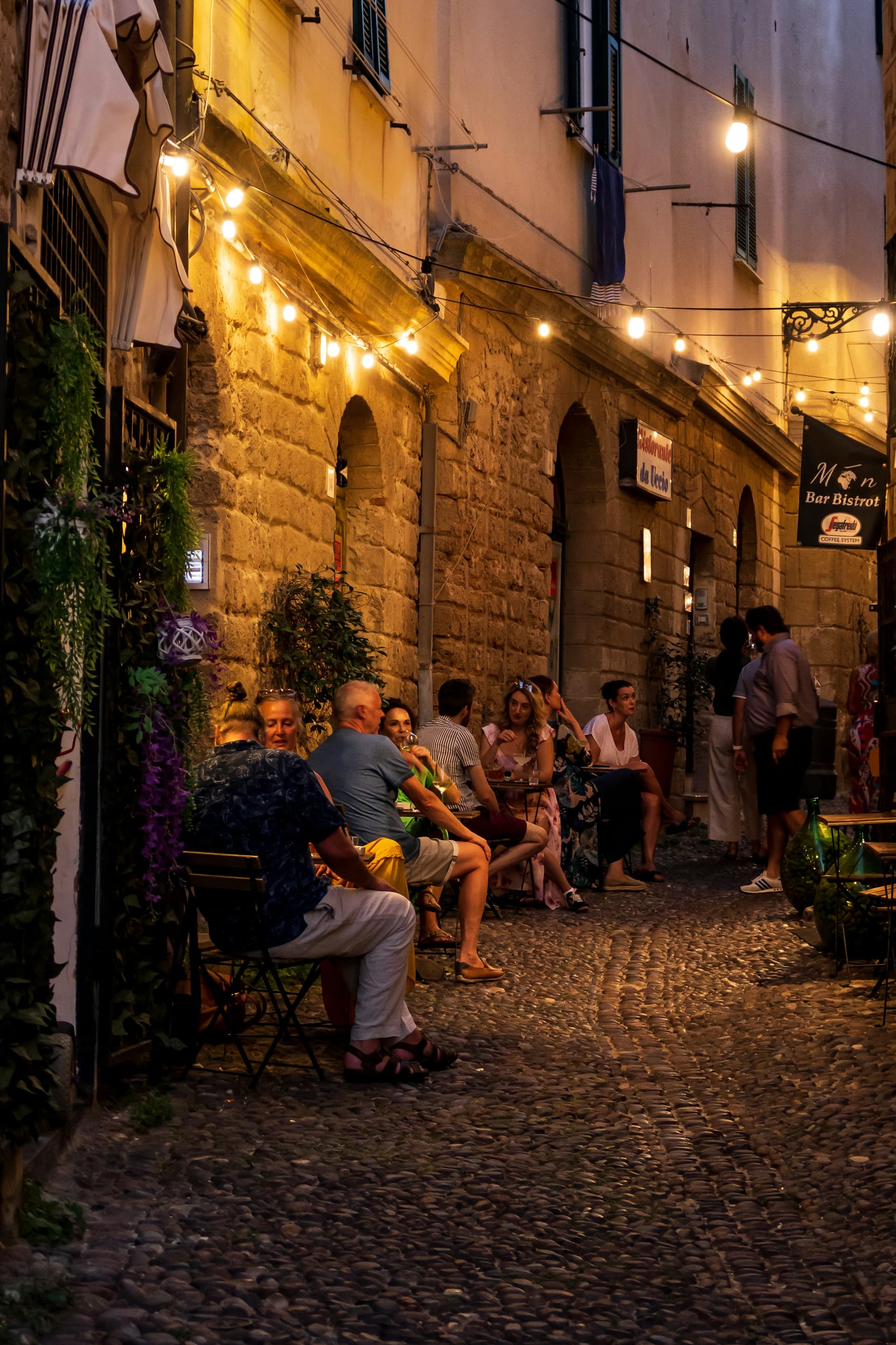Diners at tables in a cobbled lane of Alghero's old town, lit by strings of bulbs