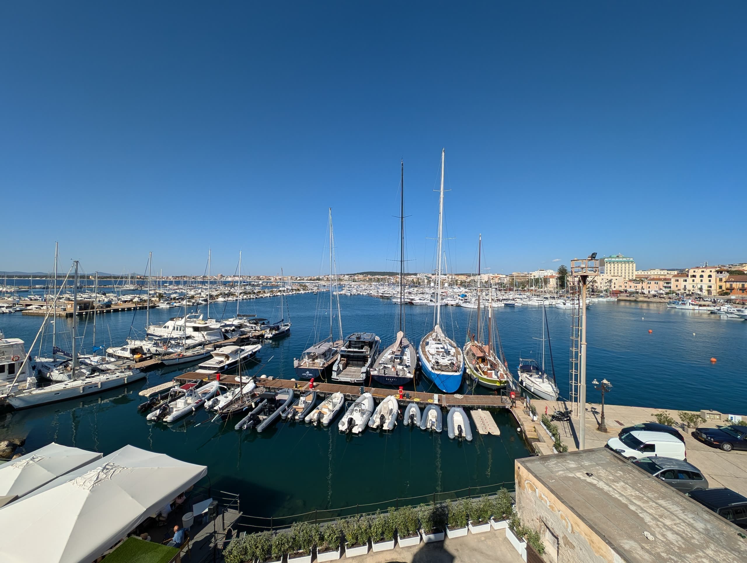 Yachts and inflatables moored in Alghero's marina, with the town's waterfront behind them