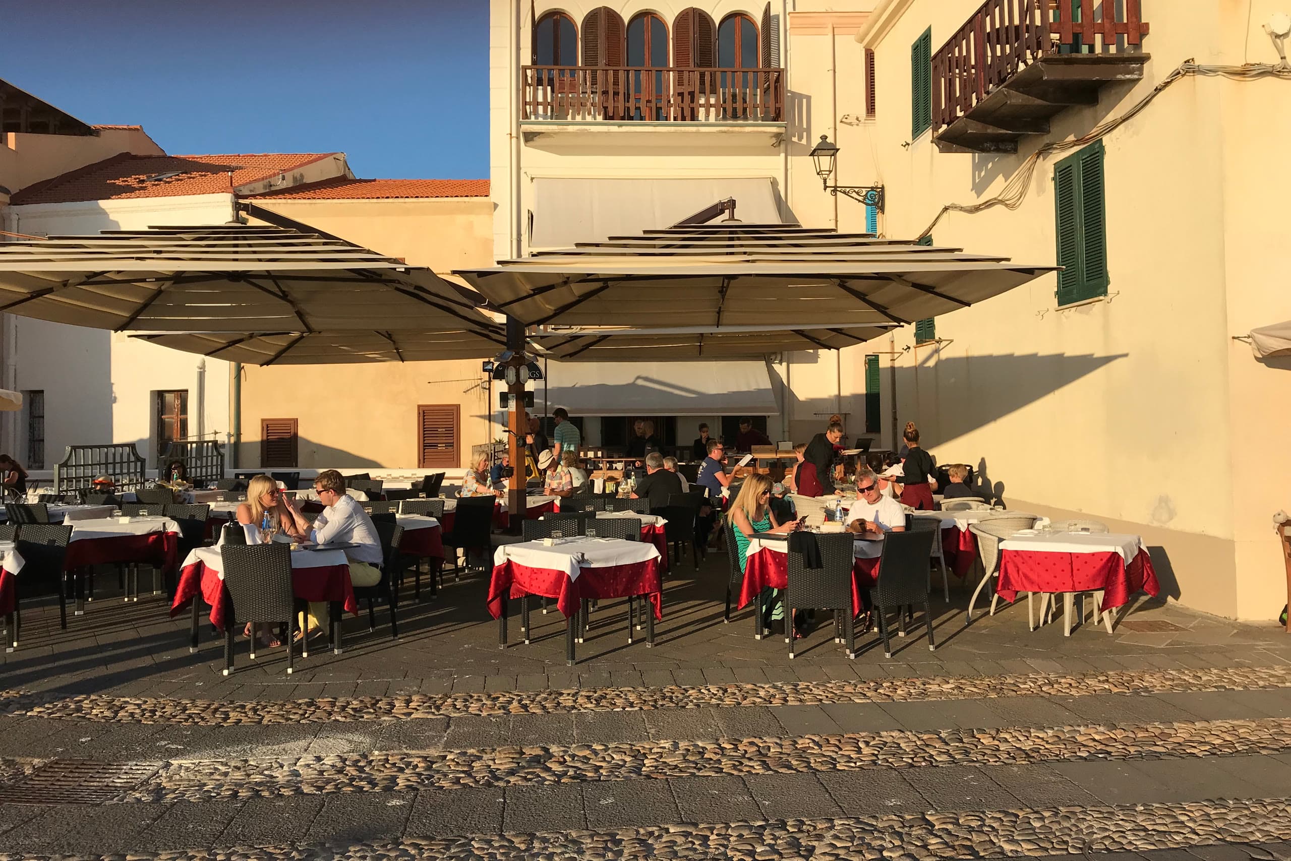 Tables laid with red and white cloths on the cobbles of Alghero's old town, full of diners in late afternoon light