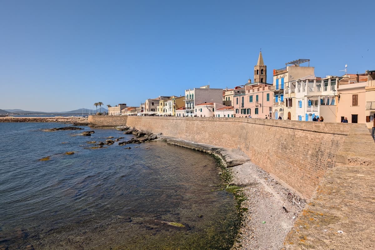 The sea bastions of Alghero above the rocks, with the old town and the cathedral bell tower behind them