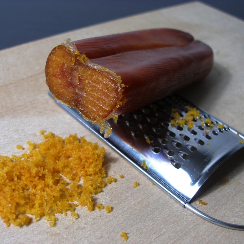 A block of mullet bottarga being grated, orange shavings collecting on the board