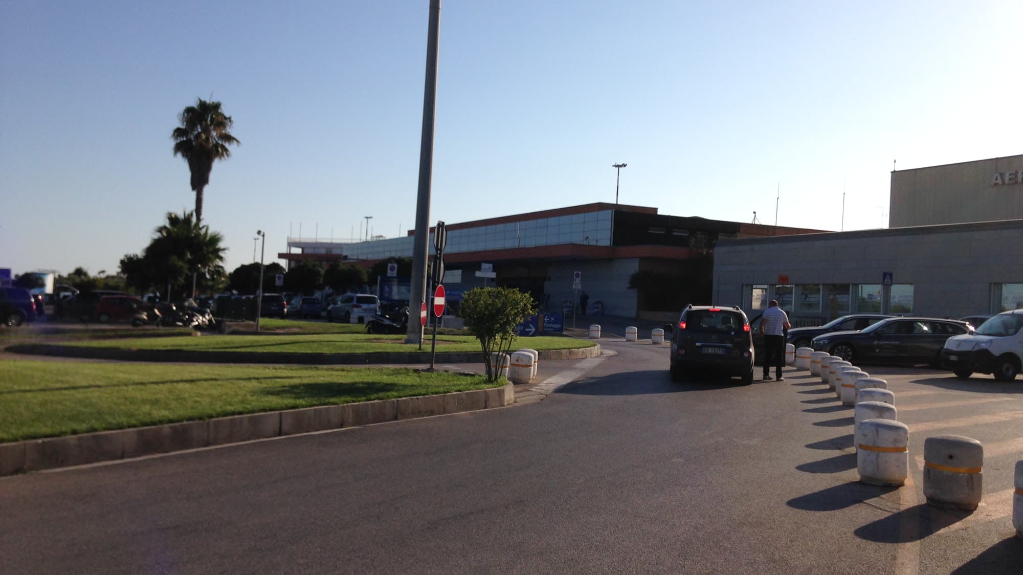 The kerbside entrance to Alghero–Fertilia airport: one low terminal building, a palm tree and the drop-off lane in front of it