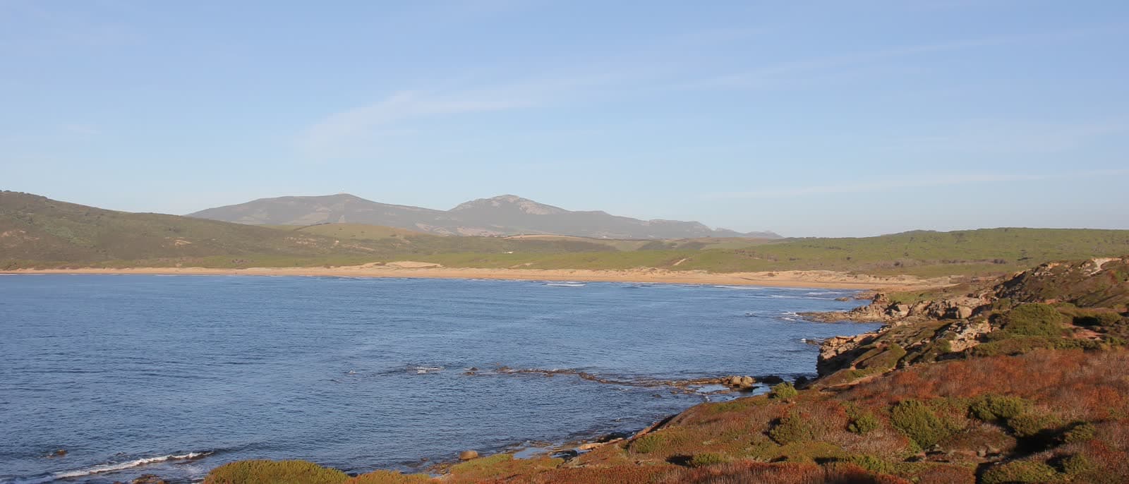 The wide bay of Porto Ferro with rust-coloured sand and dunes behind it.