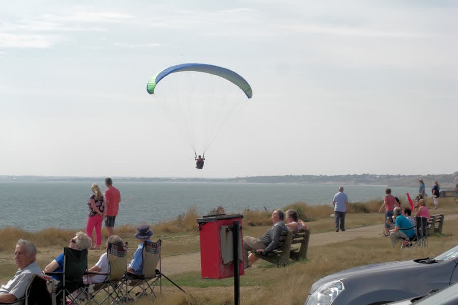 A paraglider flying along a coastal cliff, with people watching from the grass.