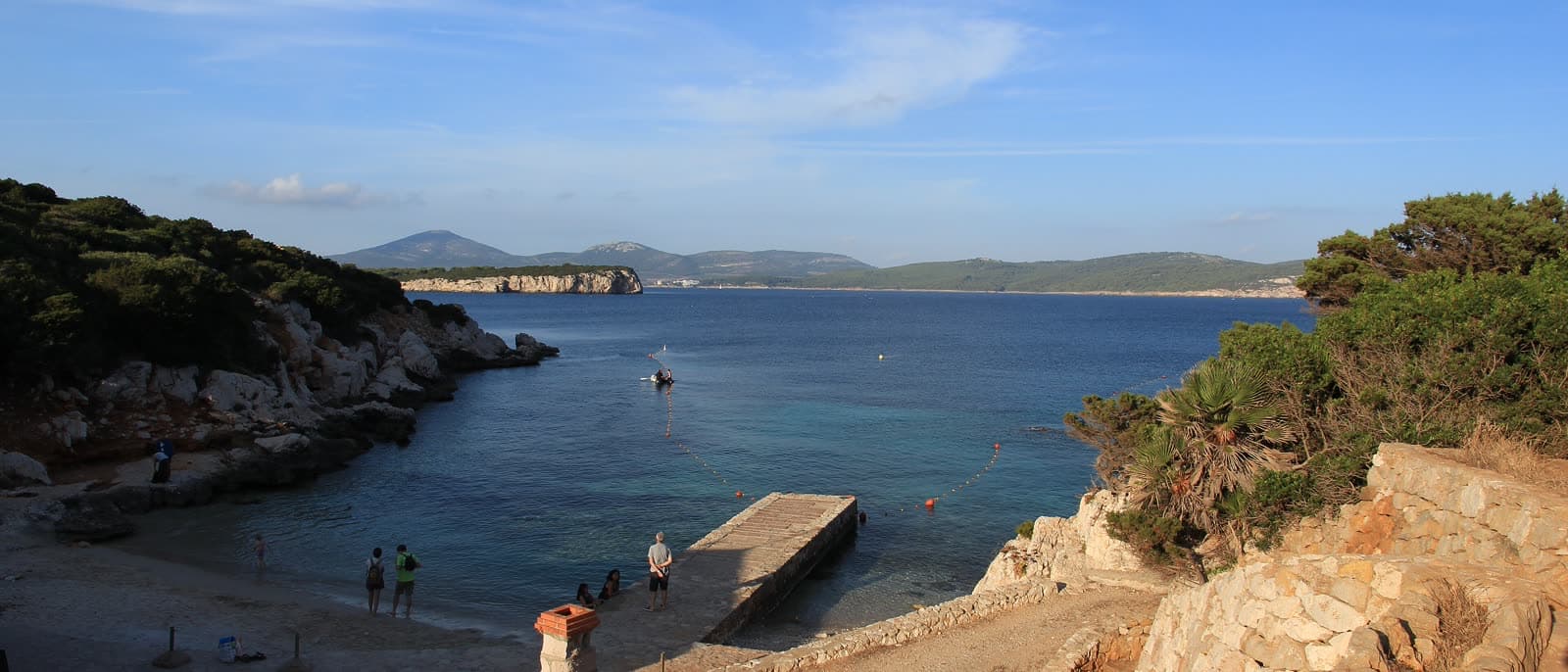 The small beach and stone jetty at Cala Dragunara, people standing at the water's edge and a sheltered bay behind.