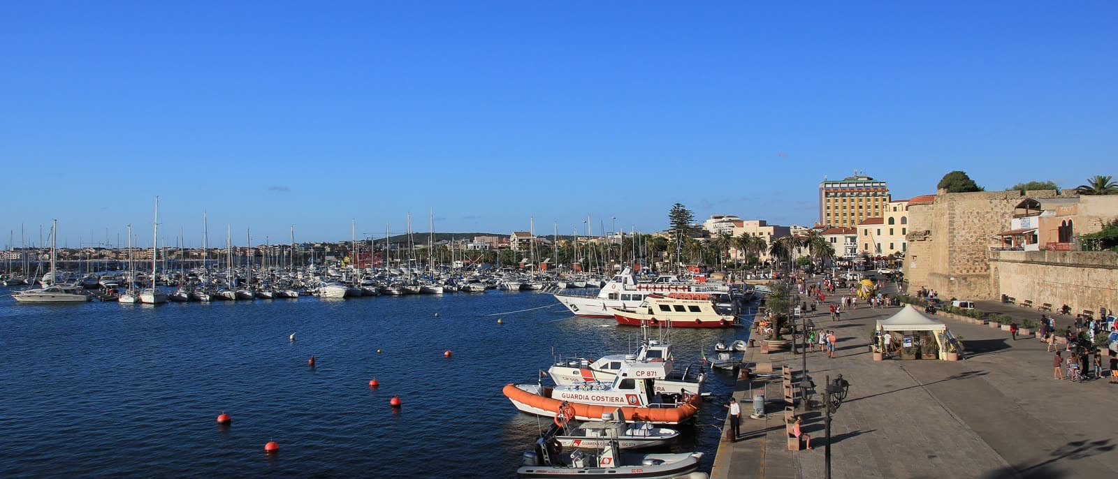 Excursion boats moored along the quay in Alghero's port, with the old town and its sea walls behind them.