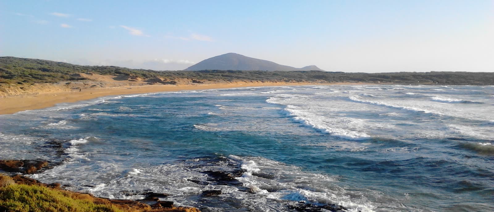 Waves rolling into the bay at Porto Ferro under a winter sky, with a low headland closing the far side.