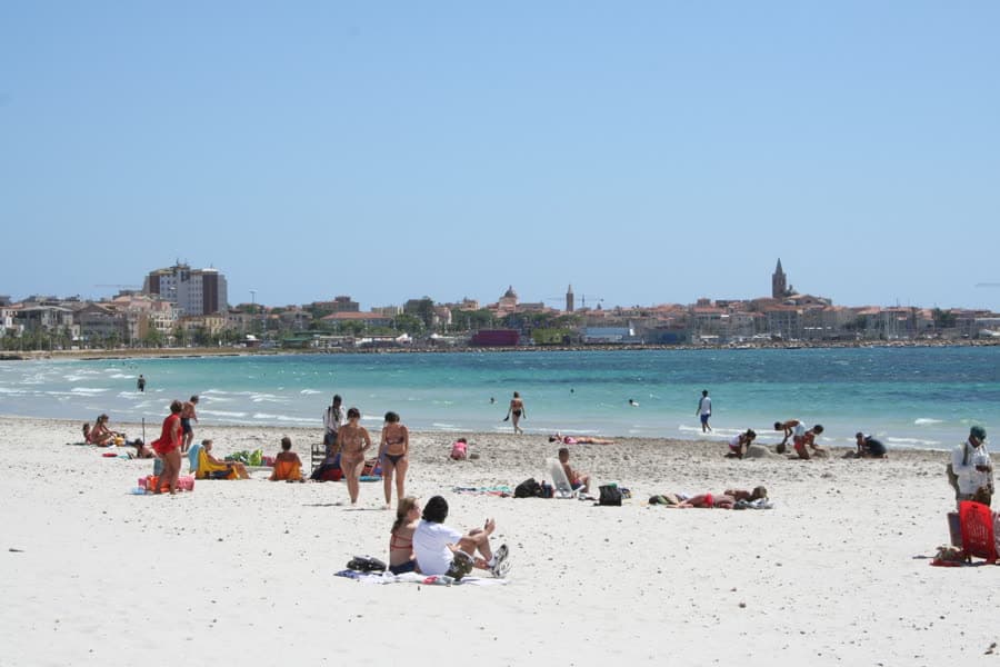 The town beach at Alghero, bathers on the sand and the old town skyline behind.