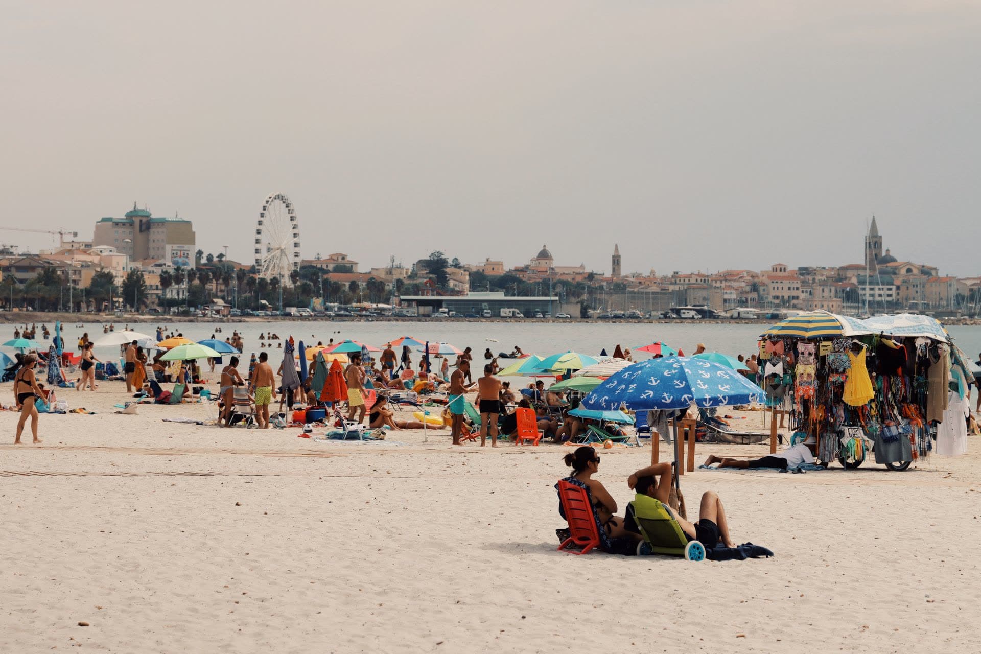 La playa urbana de Alghero un día de verano: arena blanca, sombrillas y bañistas, con el casco antiguo y sus campanarios al otro lado de la bahía