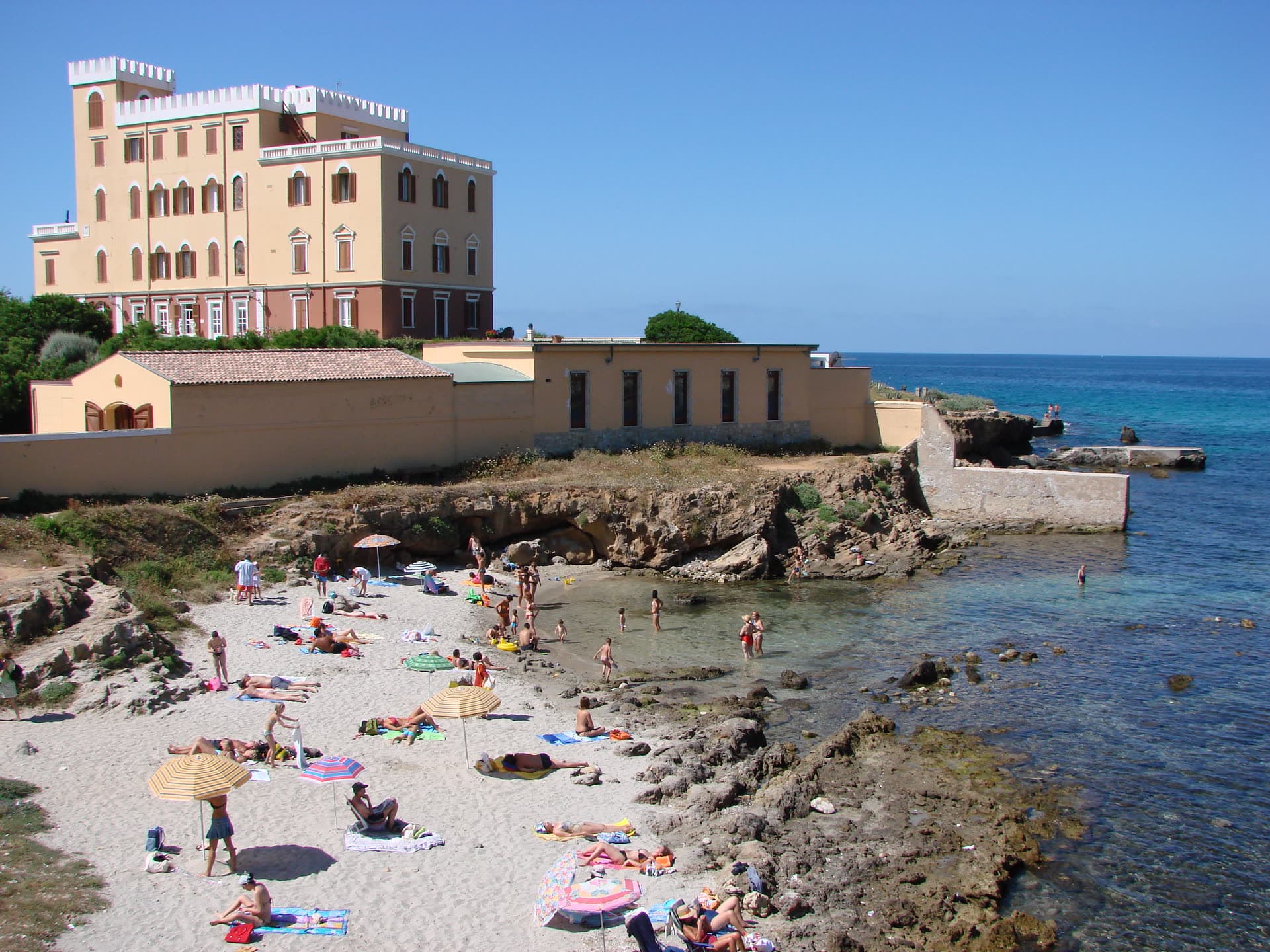 Sombrillas y bañistas en la pequeña cala de arena bajo Villa Las Tronas en Alghero, con losas de roca que entran en el agua clara