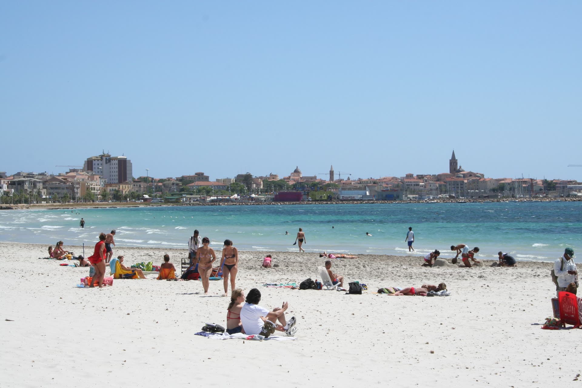 Bañistas en la ancha arena del Lido de Alghero, con el casco antiguo y el puerto deportivo al otro lado de la bahía